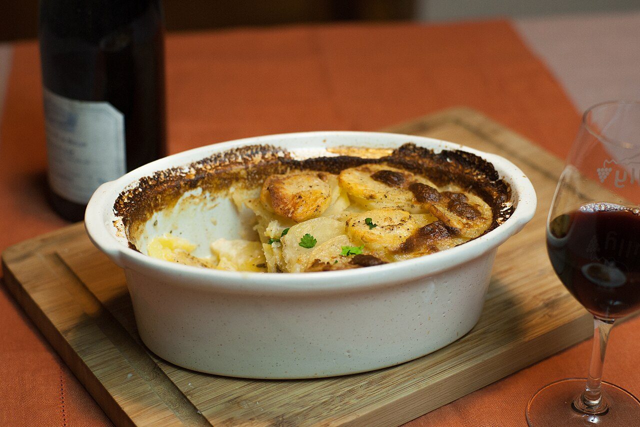 Travessa de gratin dauphinois com a superfície dourada, ao lado de uma taça de vinho tinto