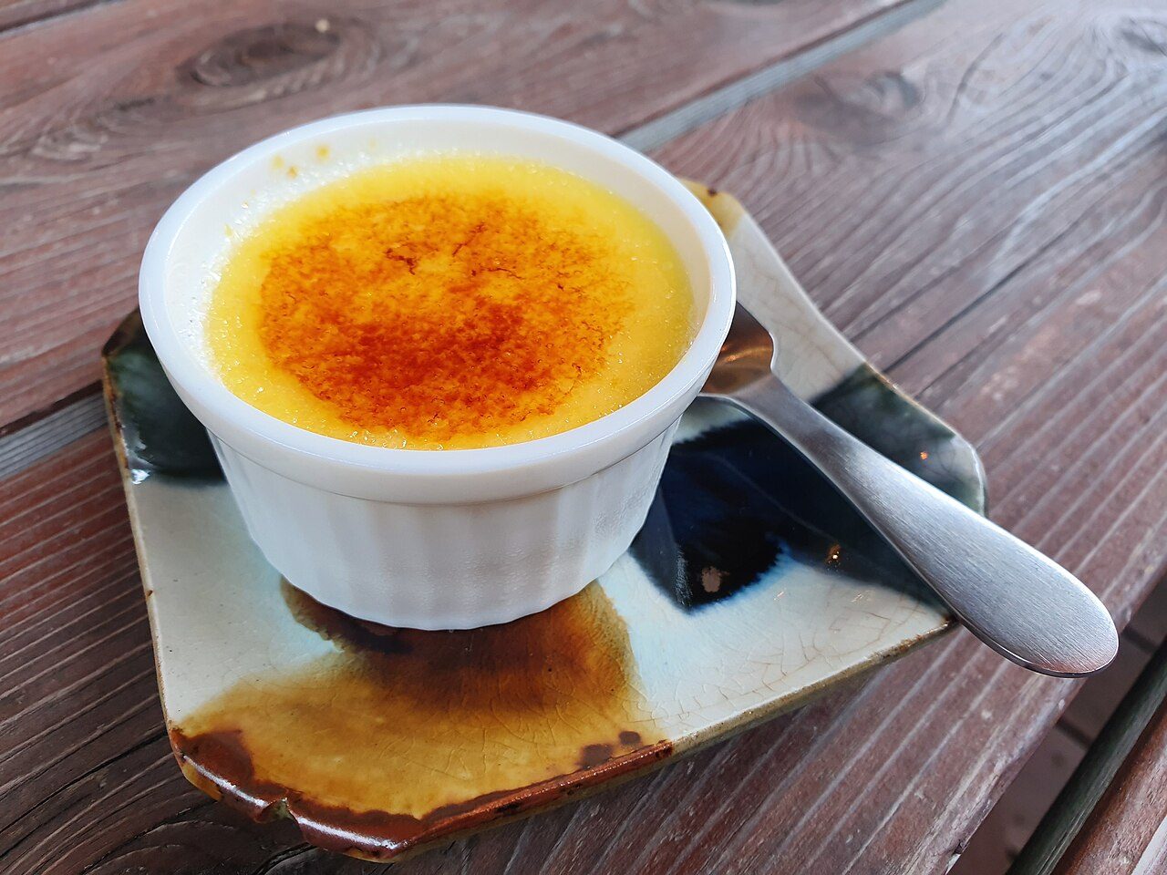 Creme brûlée em ramequim branco, com a casca de açúcar caramelizado, e uma colher ao lado