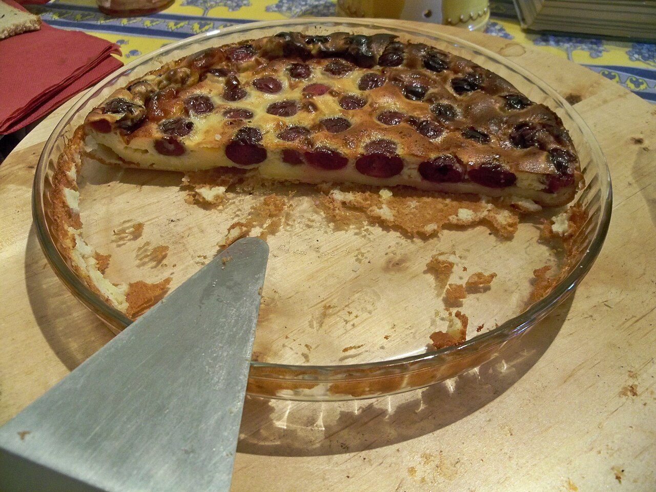 Clafoutis de cerejas num prato de vidro, já com metade servida e uma espátula à frente, mostrando as cerejas escuras inteiras presas num creme assado dourado
