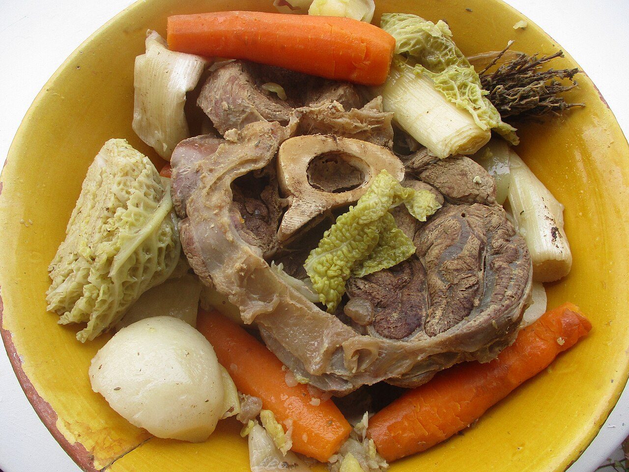 Pot-au-feu num prato amarelo, com carne, osso com tutano, cenoura, nabo, alho-poró e repolho