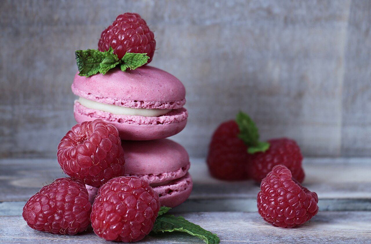 Macarons cor-de-rosa empilhados com recheio branco, mostrando a saia rendada, o pé, entre framboesas e folhas de hortelã