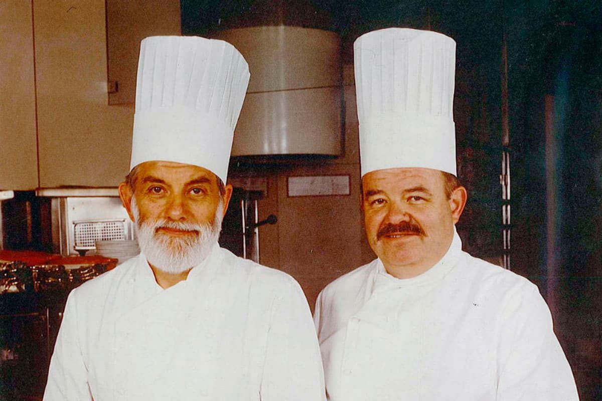Os irmãos Jean e Pierre Troisgros de dólmã branca e chapéu de chef na cozinha