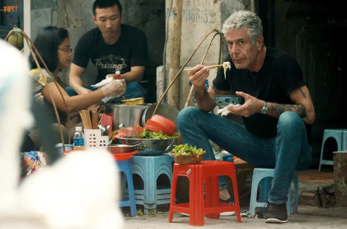 Anthony Bourdain comendo macarrão com hashi num banquinho de plástico em barraca de rua
